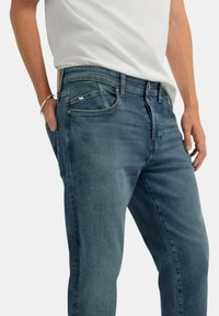 Jeans in denim blu con taglio dritto, cinque tasche e dettagli di cucitura sottili. Indossati da una persona in piedi con le mani in tasca.