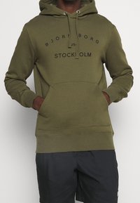 Sweat à capuche vert olive en tissu doux, avec une poche kangourou, un cordon de serrage réglable sur la capuche et un logo imprimé à l'avant.