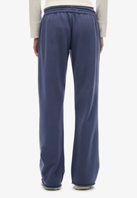 Persona con pantalones de forro polar azul marino holgados y zapatos beige, de pie contra un fondo blanco liso, vista desde atrás.