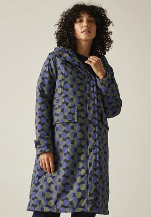 Kobieta o kręconych włosach, ubrana w długi płaszcz z kapturem w niebiesko-zielone wzory w kształcie kół, stoi na jednolitym beżowym tle.