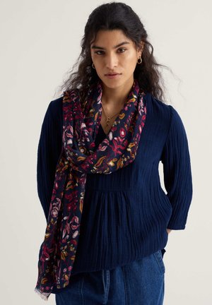 Vrouw met donker golvend haar, draagt gouden oorbellen in hoepelvorm, een marineblauwe blouse met textuur, spijkerbroek en een donker sjaal met bloemenpatroon tegen een effen achtergrond.