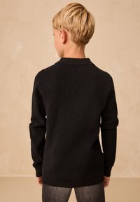 Pull noir en maille texturée avec col, manches longues et ourlet arrière légèrement plus long. Vue de dos, porté avec un jean foncé.