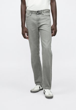 Mann trägt hellgraue Jeans mit geradem Schnitt, weiße Sneakers mit schwarzen Streifen und ein weißes Hemd vor einem schlichten Hintergrund.