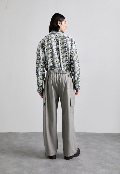 Gemustertes Hemd in Grün mit Augensymbolen; graue weit geschnittene Hose mit Seitentaschen; schwarze klobige Schuhe. Minimalistisches, zeitgenössisches Design.