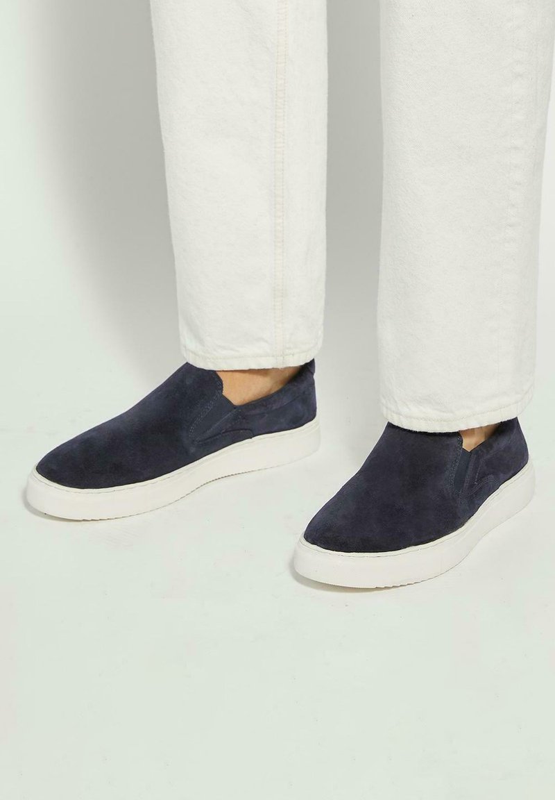 Navy blue suede slip-on shoes with a white rubber sole and elastic side accents, paired with white pants, showing smooth texture and casual design.
