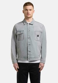 Jeune homme portant une veste en denim gris clair à boutons avec deux poches poitrine et un pantalon noir, se tenant devant un fond blanc.
