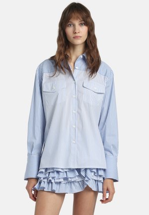 Jonge vrouw met lang bruin haar, gekleed in een blauw-wit gestreept oversized blousehemd en een bijpassend minirokje met ruches.