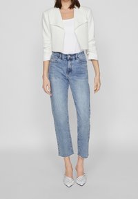 Veste blanche courte, haut blanc, jean taille haute bleu clair avec ourlets effilochés, et escarpins blancs à bouts pointus cloutés.