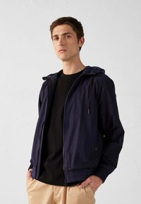 Giacca leggera blu navy con cappuccio, chiusura con zip, polsini elasticizzati e tasche laterali. Indossata sopra una camicia nera e pantaloni beige, vestibilità casual.