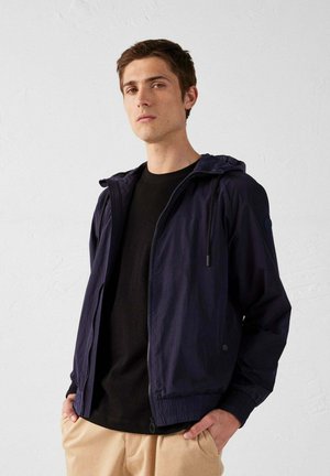 Marineblauwe lichtgewicht jas met een capuchon, ritsluiting, elastische manchetten en zijzakken. Draagt over een zwart shirt en beigekleurige broek, casual pasvorm.