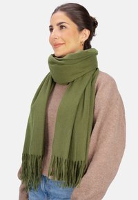 Écharpe en laine vert olive avec une texture douce, avec des franges aux extrémités, enroulée solidement autour du cou, complétant un pull beige.