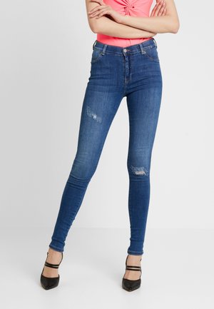 Jean skinny en denim bleu avec des déchirures aux genoux, taille haute et décoloration subtile. Associé à des chaussures à talons hauts noires et un haut rose.