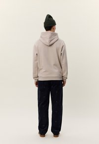 Person mit dem Rücken zur Kamera, trägt beige Kapuzenpullover, dunkel gemusterte Hose, schwarze Mütze und braune Schuhe vor schlichtem hellem Hintergrund.