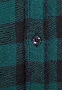 Camisa de franela a cuadros en verde y negro con un botón negro cerca de la tapeta. Tejido texturizado con patrón de cuadros y costuras visibles.