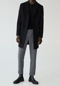 Schwarzer Wollmantel mit einreihigem Design, kombiniert mit grauen maßgeschneiderten Hosen und schwarzen Leder-Chelsea-Boots. Glatte Texturen sind sichtbar.