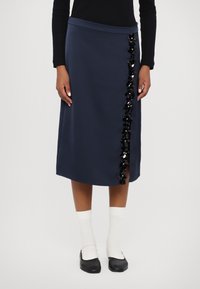 Jūras laivu zilās midi svārki ar taisnu griezumu, dekorēti ar melnām sekvīnu ziedu akcentiem kreisajā pusē un delikātu sānu šķēlumu.