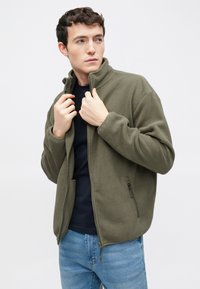 Νέος άνδρας που φοράει ένα χακί φλις μπουφάν πάνω από μια μαύρη μπλούζα και μπλε τζιν, προσαρμόζοντας το γιακά του μπουφάν, κοιτάζοντας στο πλάι.