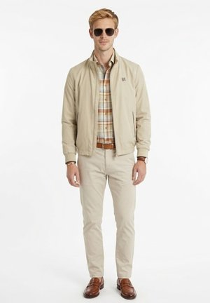 Hombre con chaqueta beige, camisa de cuadros, pantalones claros, cinturón y zapatos marrones, gafas de sol y reloj, de pie frente a un fondo blanco.