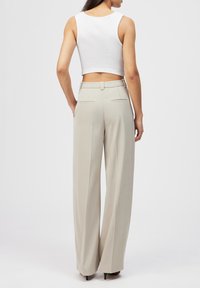 Femme portant un pantalon beige à jambes larges et un haut court blanc sans manches, debout de dos face à l'appareil photo.