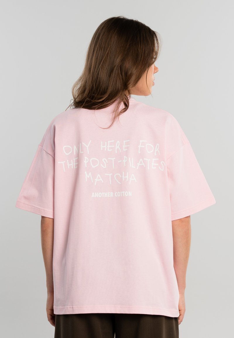 Mujer con cabello castaño que lleva una camiseta rosa holgada con texto blanco en la parte posterior, sobre un fondo gris liso.