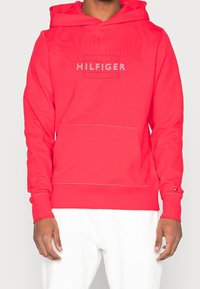 Red hoodie with a front kangaroo pocket, "HILFIGER" logo in white, ribbed cuffs and hem, made from soft material.