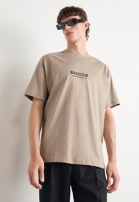 Beige puuvillane T-särk lühikeste varrukatega ja ümmarguse kaelusega, mille esiküljel on musta värviga trükitud "BARBOUR INTERNATIONAL". Kandmine mustade pükstega.