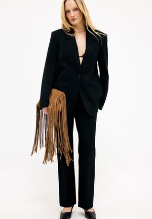 Žena v čiernom šitom obleku na mieru a čiernych lodičkách drží veľké hnedé semišové psaníčko s dlhými strapcami na bielom pozadí.