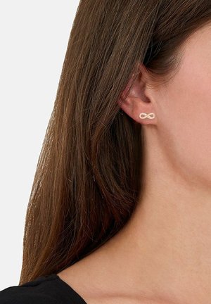 Orecchini a punto oro a forma di simbolo dell'infinito, con finitura liscia e dettagli subtili. Indossati all'orecchio di un modello.