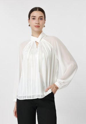 Vrouw draagt een witte doorschijnende blouse met lange mouwen en een hoge hals, gecombineerd met een zwarte broek. Ze poseert met één hand in de zak en heeft een neutrale gezichtsuitdrukking.