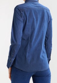 Jeansskjorta i mörkblått, långa ärmar, knäppning vid ärmslut, strukturerad ryggsömn, och en något rundad nederkant.