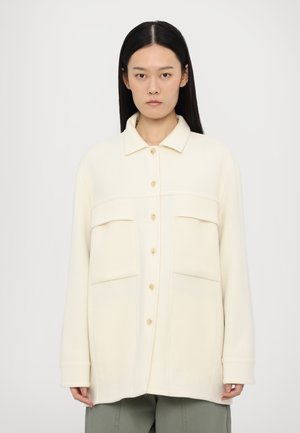 Crème-kleurige oversized shirtjas gemaakt van zachte stof, met een kraag, een knoopsluiting aan de voorkant en twee grote opgestikte zakken.