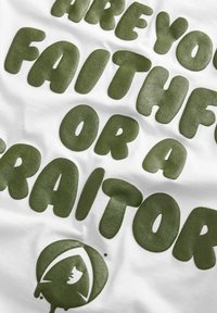 Hvid bomulds t-shirt med store grønne teksturerede bogstaver, der står "ER DU TROFAST ELLER EN FORRÆDER?" med et rundt logo i bunden.