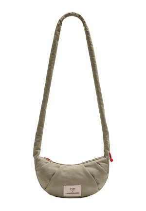 Kleine beigenfarbene Schultertasche in Halbmondform mit langem gepolstertem Träger und rotem Reißverschlussanhänger, mit einem weißen Etikett mit schwarzem und rotem Text.