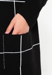 Czarny sweter z białym wzorem w kratkę, posiadający przednią kieszeń i fakturowany materiał. Ręka częściowo widoczna wewnątrz kieszeni.