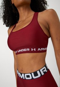 Mujer vistiendo un sujetador deportivo y leggings burdeos de Under Armour, mostrando el torso superior y la cintura con logotipos de la marca visibles en las bandas elásticas.