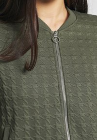 Olivgrön bomberjacka med en texturerad yta prydd med diamantmönster, metallblixtlås och ribbade muddar samt krage.