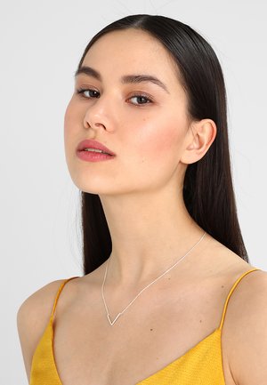 Collier en argent avec un pendentif en forme de V, porté par un modèle. Le design du pendentif est épuré, avec une fine chaîne contre un haut jaune.