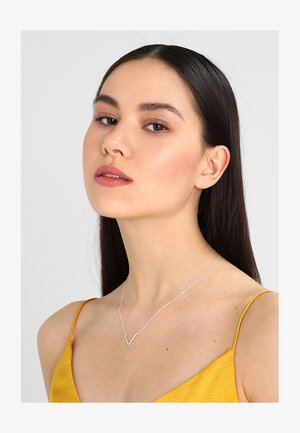 Necklace argintie cu un pandantiv în formă de V, purtată de un model. Designul pandantivului este elegant, cu un lanț subțire pe un top galben.
