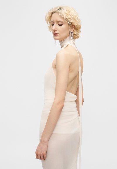Blonde woman with short curly hair wearing a backless pale dress and long crystal earrings, looking down and to the side.