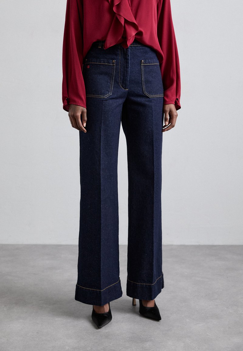 Φαρδύ τζιν παντελόνι από σκούρο ντένιμ με αντίθετη κίτρινη ραφή και μανίκια. Συνδυάζεται με μπλούζα μακρύ μανίκι σε κόκκινο χρώμα, με κρόσσια στον λαιμό.