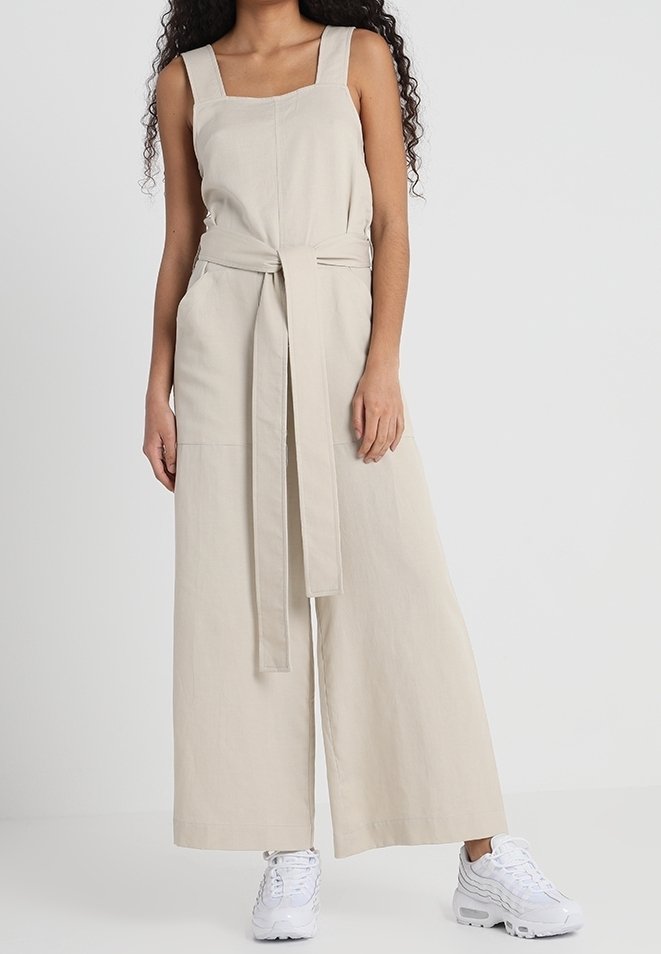 Femme portant une combinaison beige sans manches à jambes larges avec ceinture en tissu, associée à des baskets blanches sur fond uni.