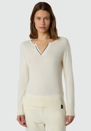 Donna che indossa un maglione a maniche lunghe di colore bianco sporco con una piccola tacca e bordo scuro al collo, abbinato a pantaloni color crema.