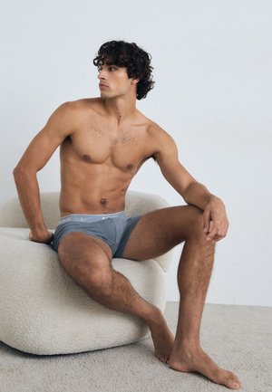Graue Nike Boxerbriefs mit einem Logo am Bund, getragen von einem sitzenden männlichen Modell auf einem strukturierten, runden weißen Stuhl.