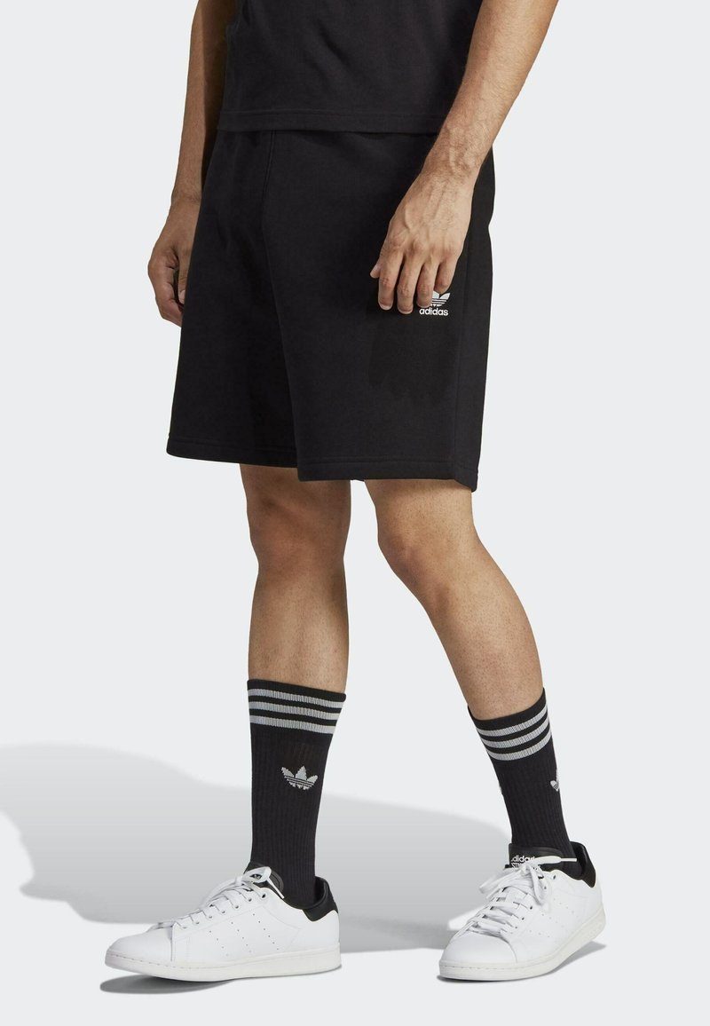 Shorts neri in cotone con tasche laterali, calze a strisce nere e grigie coordinate e sneakers bianche con accenti neri. Logo visibile sui pantaloni.
