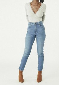 Graues Langarm-Wickeltop, hoch taillierte blaue Jeans und braune Stiefeletten. Die Jeans haben einen schmalen Schnitt; das Top hat einen gewellten Ausschnitt.