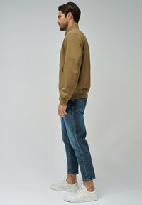 Veste bomber marron clair avec des poignets côtelés, associée à un jean en denim bleu. Des baskets blanches complètent le look décontracté. Texture lisse et design ajusté.