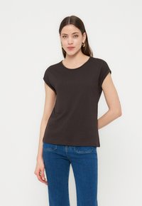 Γυναίκα που φοράει μαύρο κοντομάνικο μπλουζάκι και μπλε τζιν, όρθια με το ένα χέρι πίσω από την πλάτη, μπροστά σε ένα απλό λευκό φόντο.