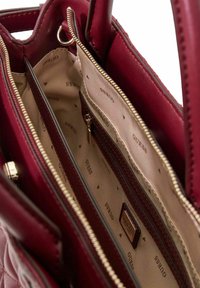 Sac à main bordeaux avec un extérieur matelassé, doté d'une doublure intérieure beige embossée de triangles et du texte "GUESS". Comprend des compartiments zippés.