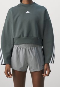 Mörkgrön kortärmad sweatshirt med en liten vit logotyp och grå träningsshorts med elastiskt midjeband och tre vita ränder på sidorna.