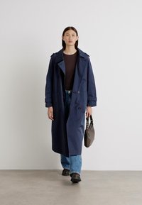 Marine trenchcoat med en avslappet passform, dobbeltradsdesign, store revers og sidelommer, kombinert med lyseblå jeans og svarte støvler.